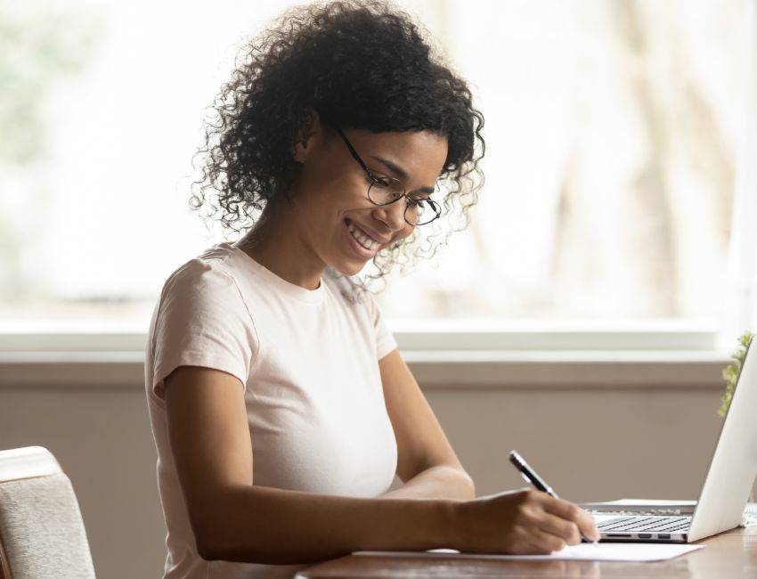 Satisfied black woman housewife in glasses sitting at table near laptop holding pen writing planning family budget making notes, businesswoman working at home, girl studying online noting information