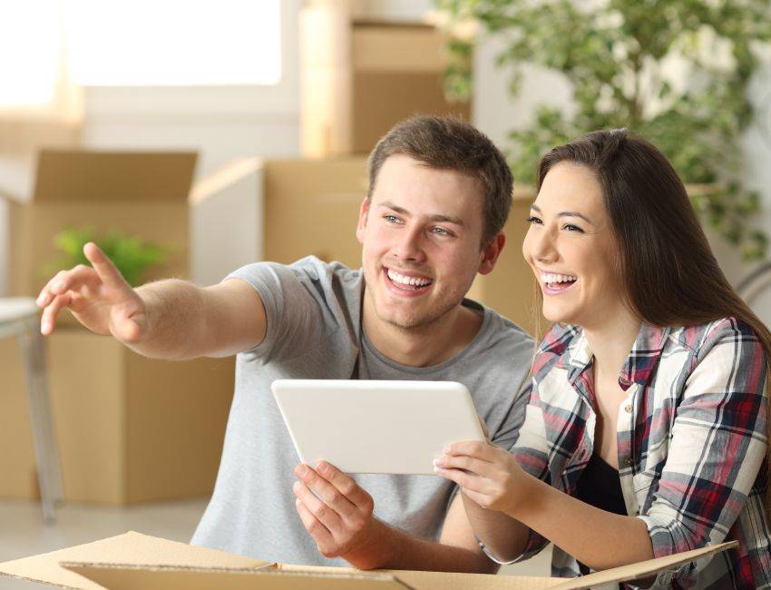 Happy couple moving house together planning on line with a tablet sitting on the floor at home with boxes in the background